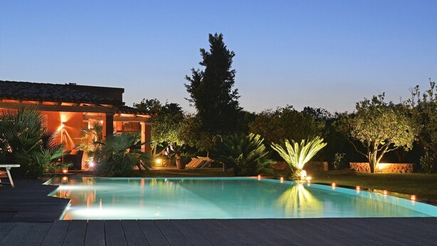 A swimming pool with a wooden deck, surrounded by trees and a building with outdoor lighting.