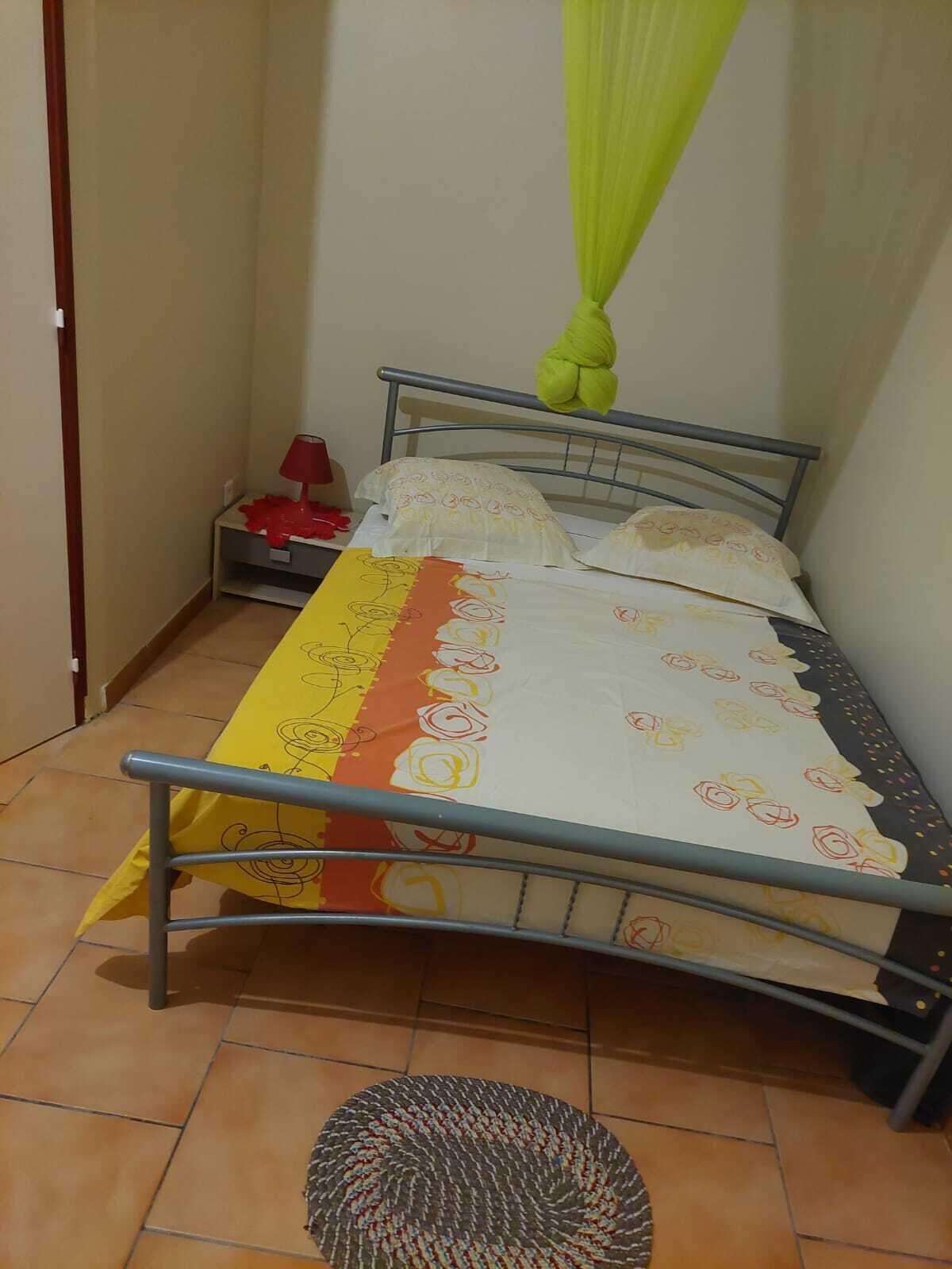 Uma cama de solteiro com estrutura de metal, edredom com padrão amarelo e laranja, e um dossel verde.
