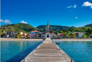 A wooden pier leading to a church with a steeple, surrounded by houses and palm trees.