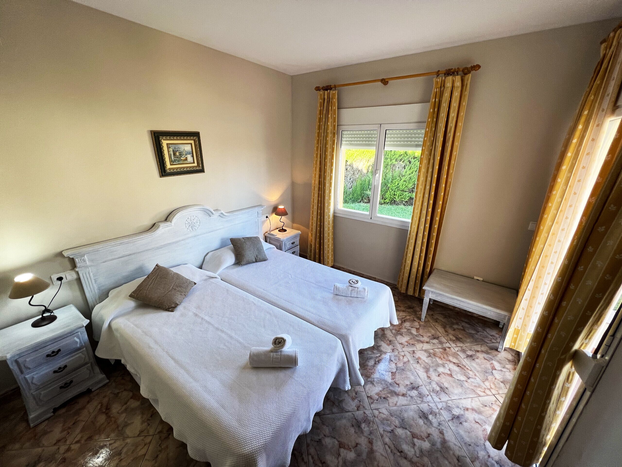 A double bed with a headboard, a bedside table with a lamp, a window with curtains, and a framed picture on the wall.