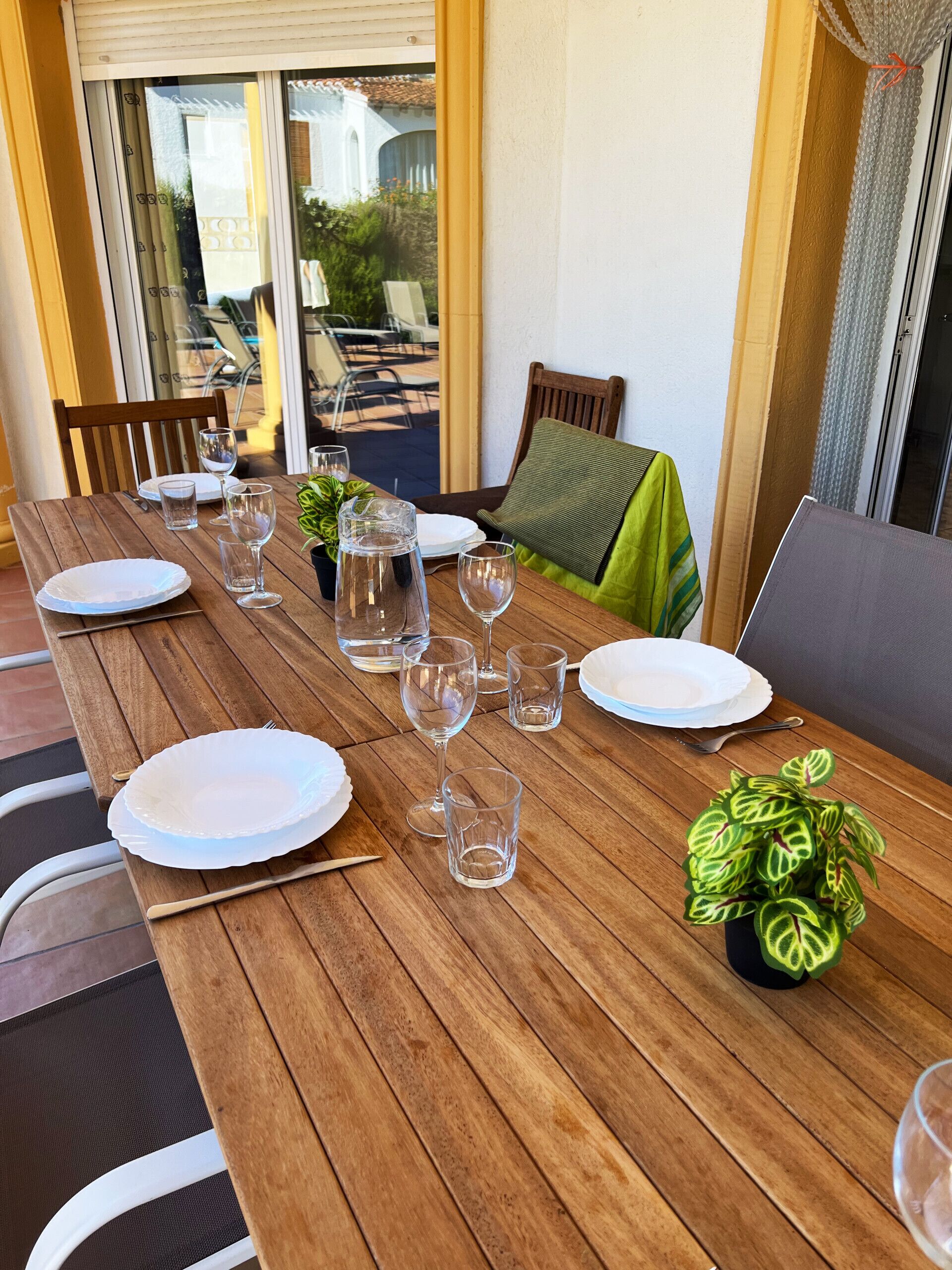 A wooden dining table set with plates, glasses, and cutlery, surrounded by chairs and a potted plant.