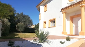 Un bâtiment blanc orné de bordures et de colonnes jaunes, un jardin bien entretenu avec du gravier blanc et des plantes, et un ciel bleu dégagé.