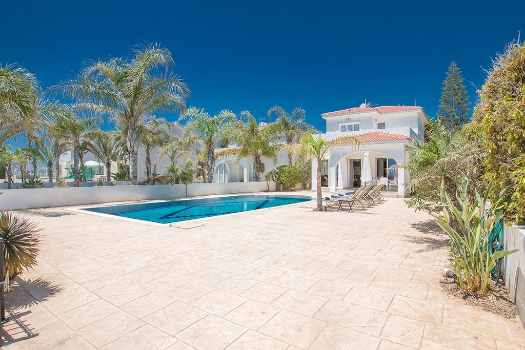 A pool area with a clear blue pool, surrounded by palm trees and a white building with a red roof.