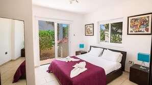 A hotel room with a large bed, a sliding glass door to the outside, a small bedside table with a lamp, and two framed artworks on the wall. - Ayia Napa Holiday Villa SV11 - Stunning 3BDR Seafront Ayia Napa Villa with Pool (Unknown)