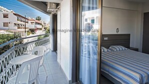A balcony with white chairs, a bed with striped bedding, and a view of neighboring buildings.