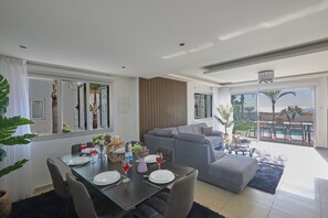 Un salon moderne avec un coin repas, des canapés gris et une grande porte-fenêtre coulissante donnant sur une piscine.