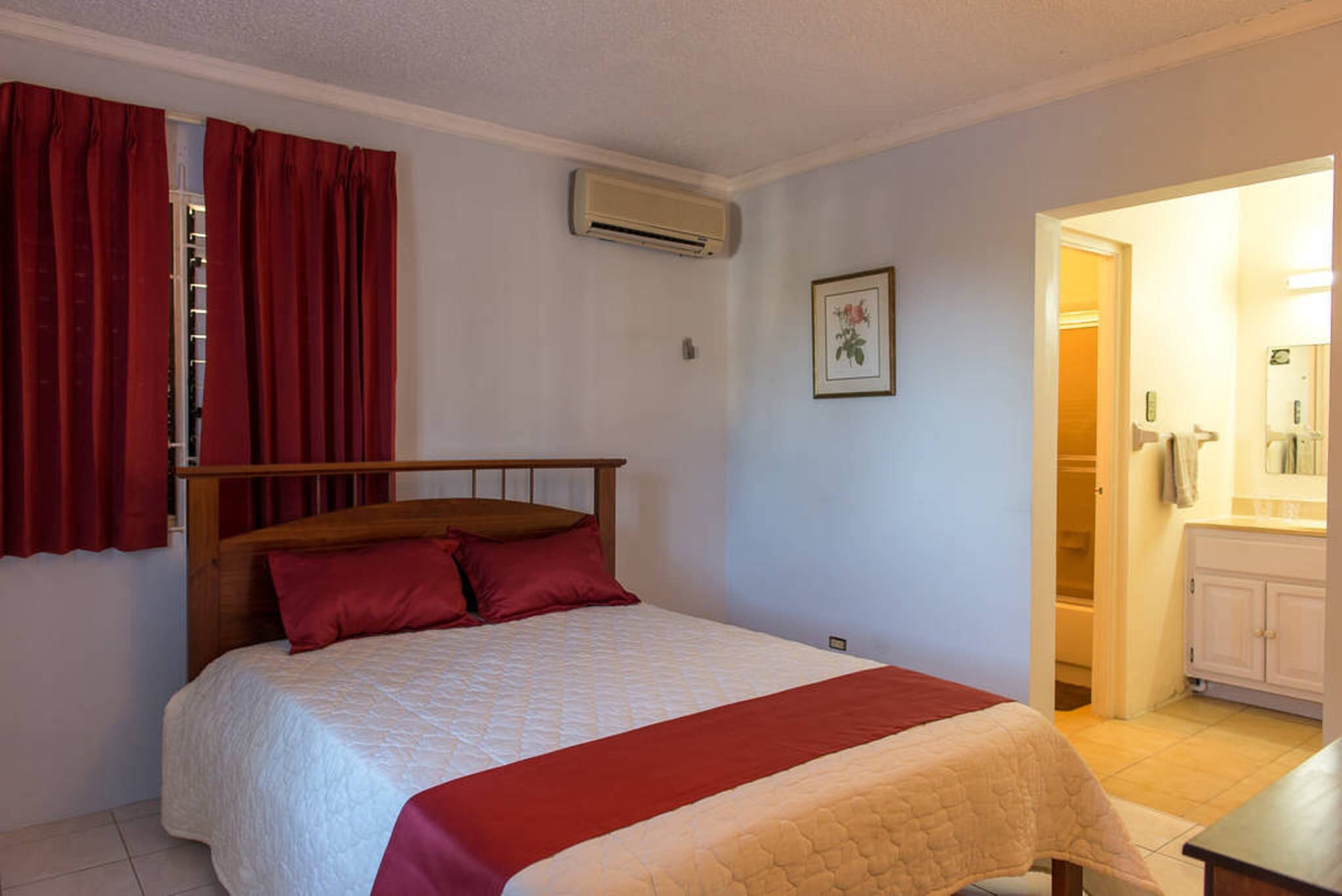 Un dormitorio con cama, cortinas rojas, un equipo de aire acondicionado, un cuadro enmarcado y un baño visible a través de una puerta abierta.