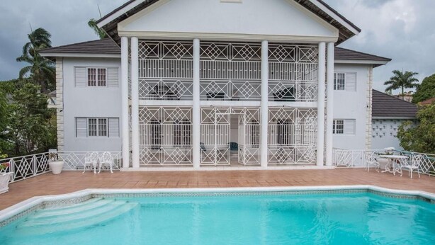 Un edificio de dos pisos con un balcón grande, piscina y mobiliario exterior blanco.