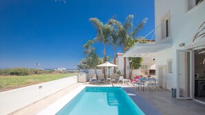 En swimmingpool med liggestole og parasoller, en patio med bord og stole, og udsigt til havet.