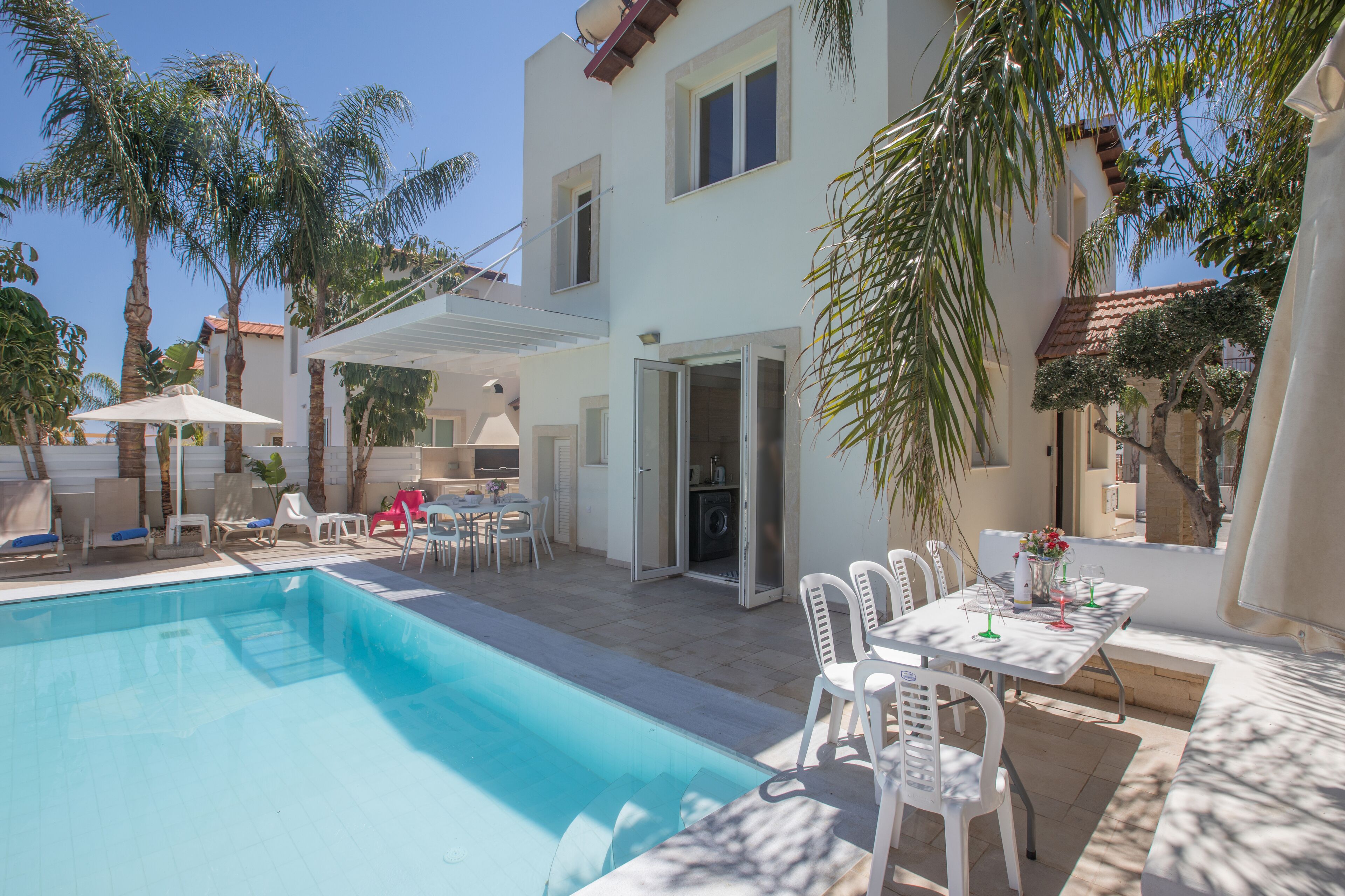 Zona junto a la piscina con piscina, sillas blancas, una mesa con un florero y palmeras.