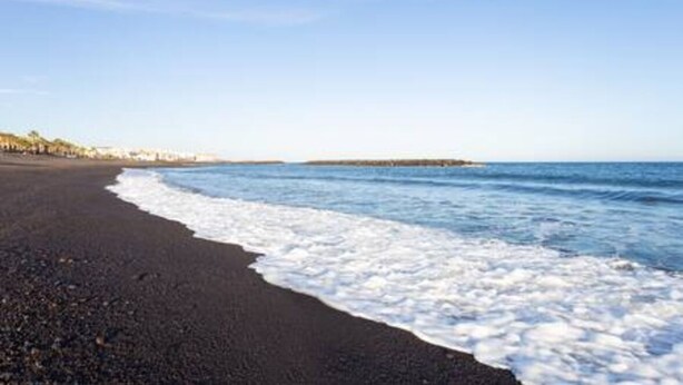 Una playa de arena oscura, con olas suaves y un cielo despejado.