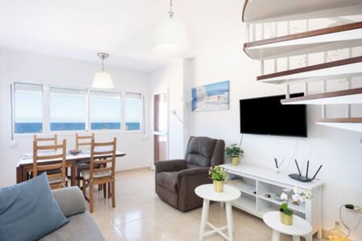 A modern living room with a sofa, a dining table with chairs, a TV mounted on the wall, and a staircase.