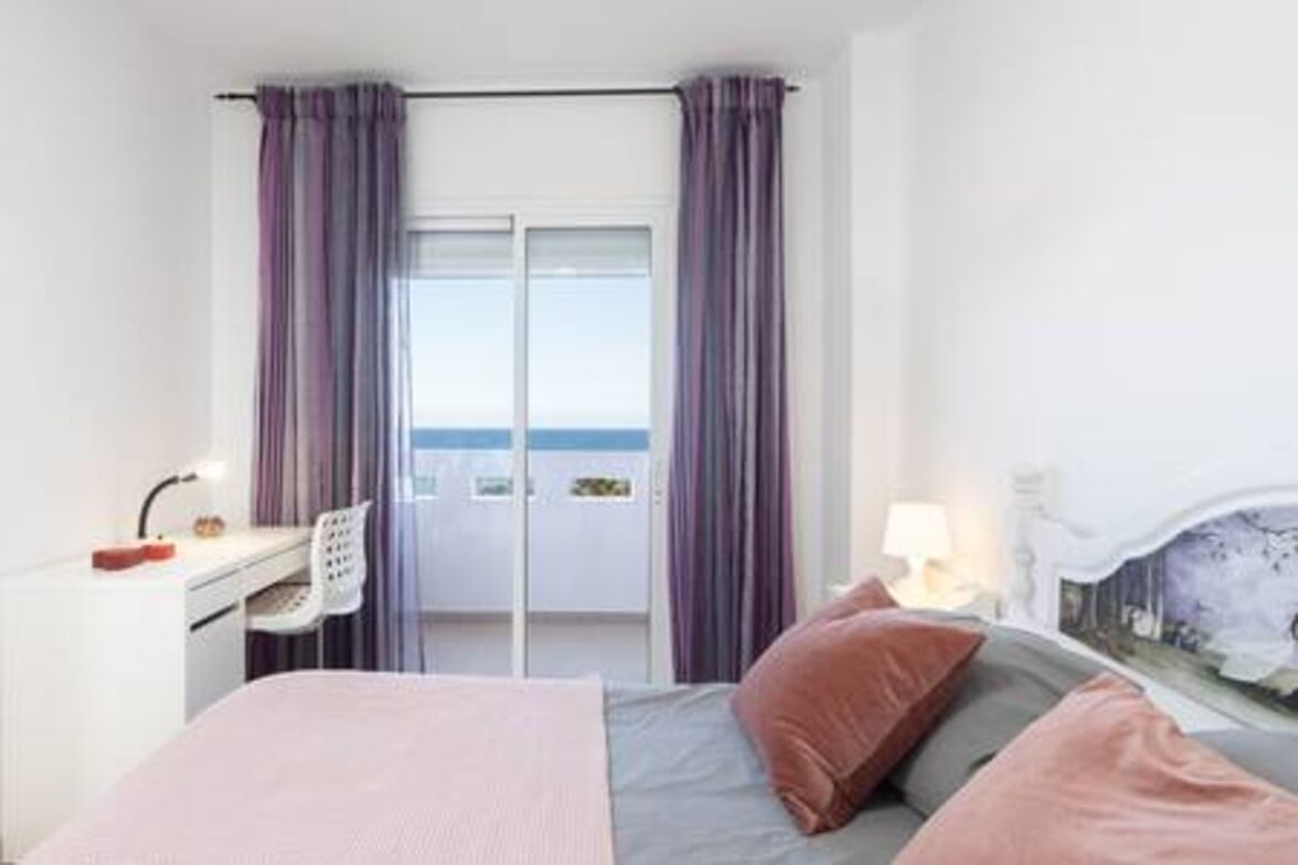 Een hotelkamer met een bed, een bureau met stoel, een raam met gordijnen en uitzicht op zee.