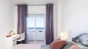 Habitación de hotel con cama, escritorio con silla, ventana con cortinas y vista al mar.