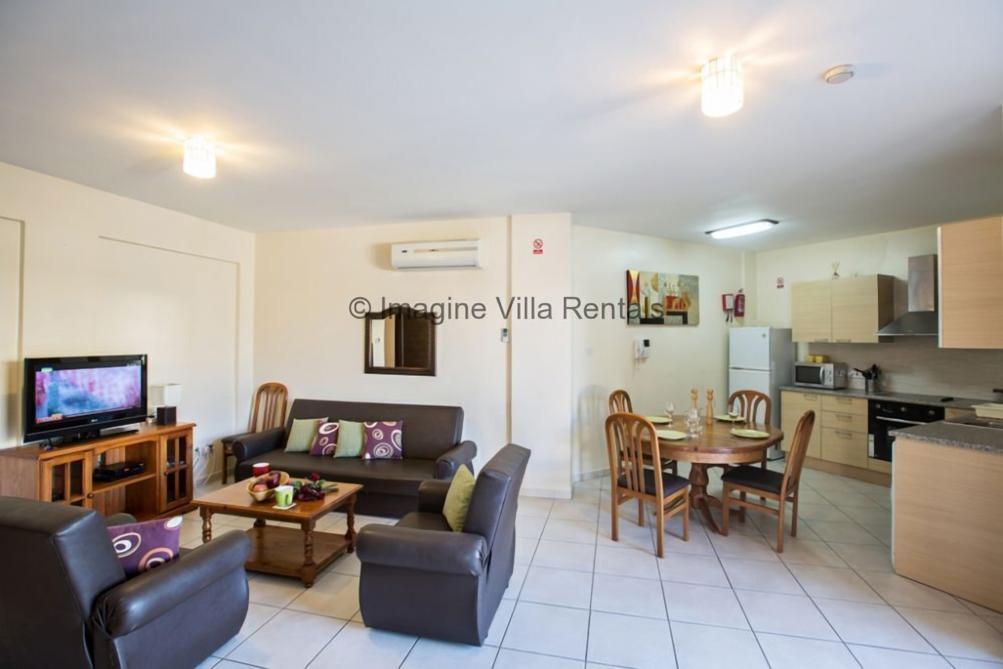 Una sala de estar con sofá, mesa de centro y televisor, un comedor con sillas y mesa, y una cocina con armarios y electrodomésticos.