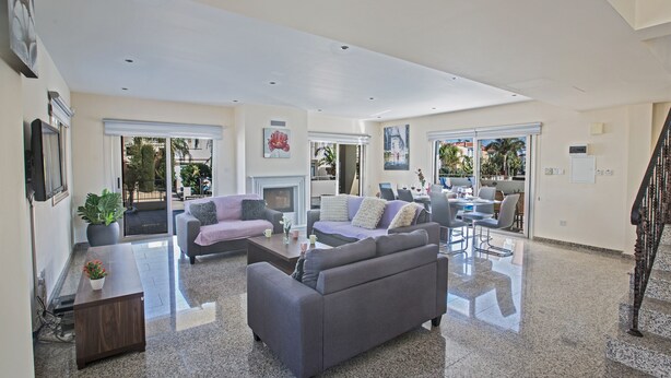 A spacious living area with a grey sofa, a coffee table, and a TV. There are large windows and a staircase leading to another level.