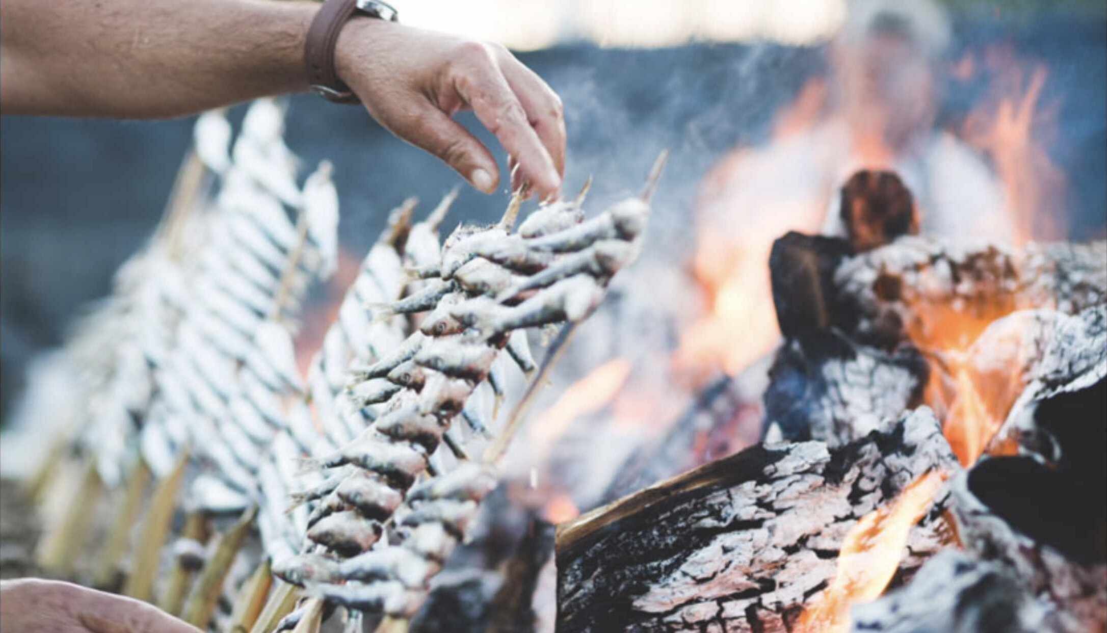A hand holding a skewer with food items over a campfire.