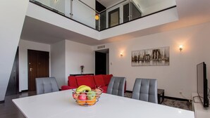 Een moderne eethoek met een witte tafel, rode en grijze stoelen en een schaal fruit. Een stadsgezichtschilderij versiert de muur, en er is een tv aan de muur bevestigd.