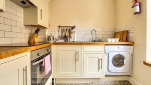 Een kleine keuken met witte kasten, een houten aanrecht, een inbouwoven, een wasmachine en een brandblusser aan de muur.