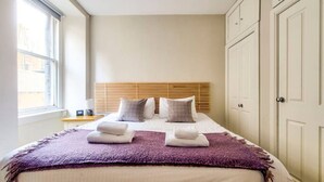 Een net opgemaakt bed met een paars plaid en twee handdoeken, een houten hoofdbord en witte kasten aan de rechterkant.