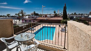 Un espace piscine aménagé sur le toit, avec une table et des chaises, entouré d’une clôture métallique, et offrant une vue sur le paysage urbain avec ses bâtiments et ses palmiers.