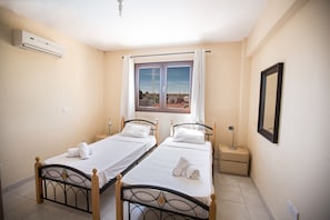A room with two single beds, a window with curtains, an air conditioning unit, and a bedside table with a lamp.