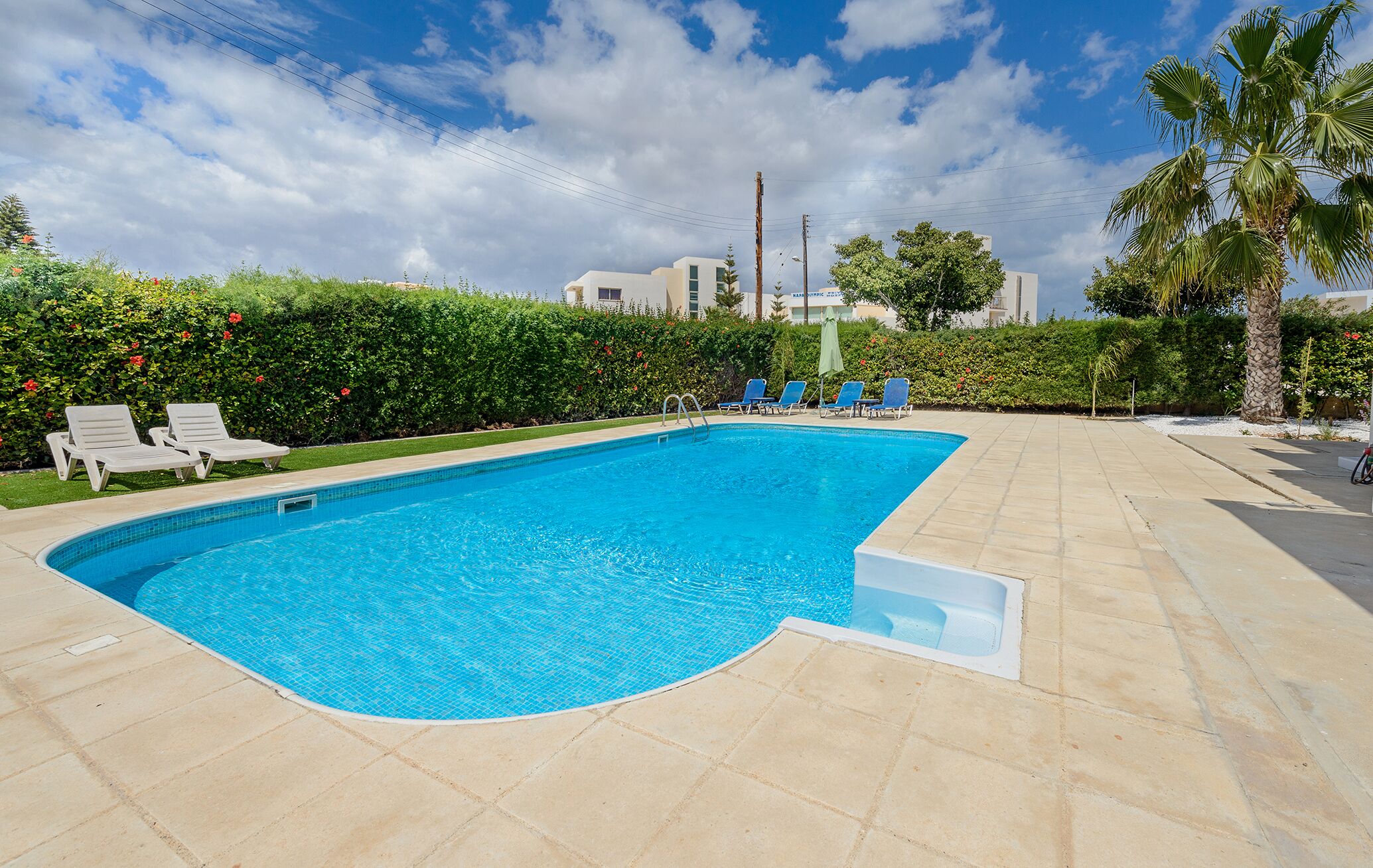 Una piscina con bordo curvo, circondata da piastrelle color beige. Sono presenti diverse poltrone prendisole blu e un piccolo ombrellone.