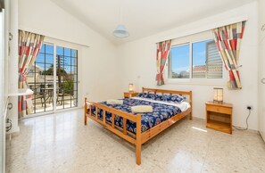 Un dormitorio con cama de madera, mesita de noche, ventana con cortinas y balcón con vistas a edificios.