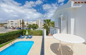 Zona de piscina con sillas de descanso, una mesa y vistas a edificios al fondo.