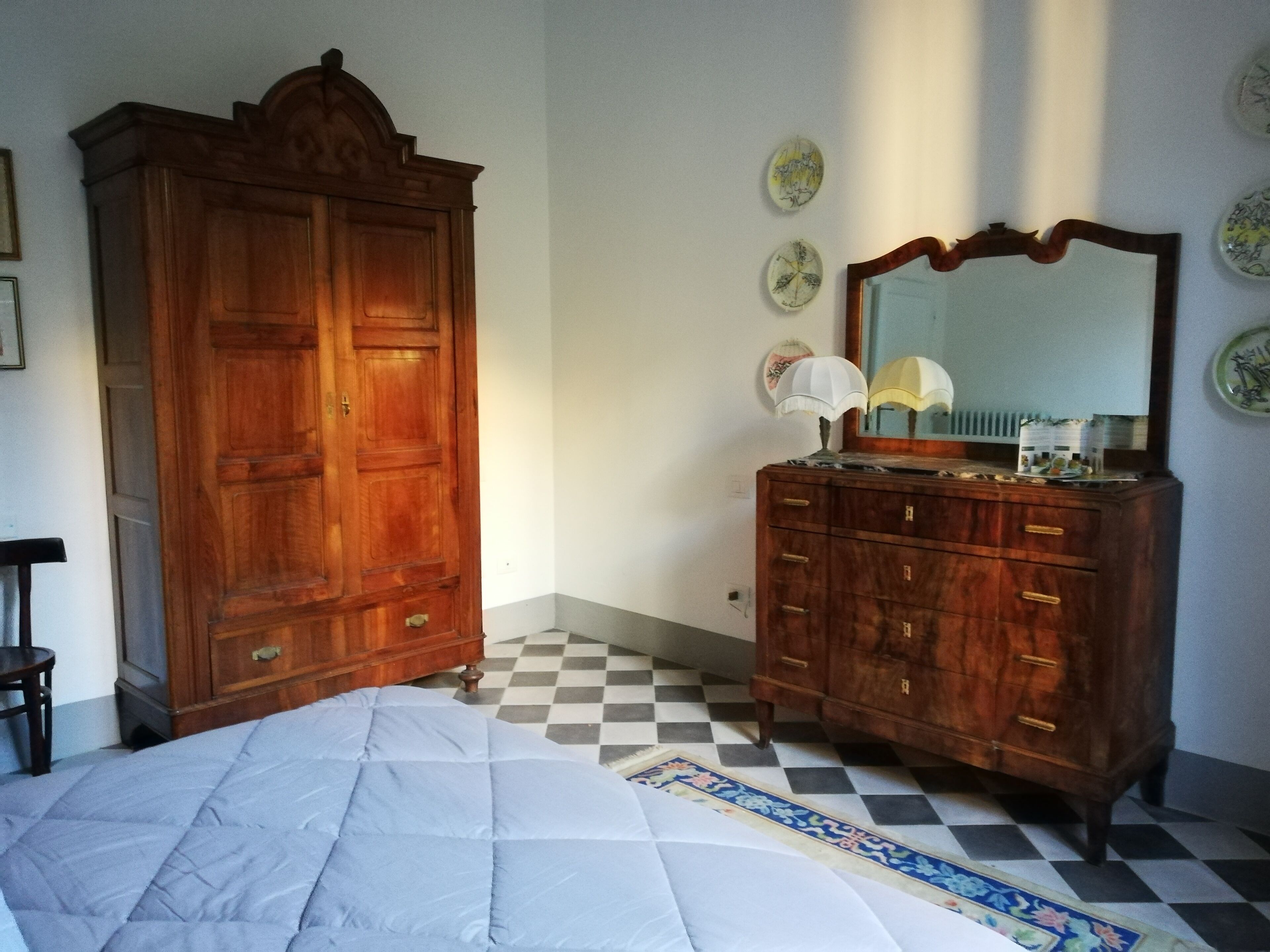 Een slaapkamer met een grote houten garderobe, een bed met quilt, een kaptafel met spiegel en een geruit vloerkleed.