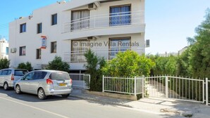 Un edifici blanc de dos pisos amb balcons, una entrada tancada amb reixa i cotxes aparcats.
