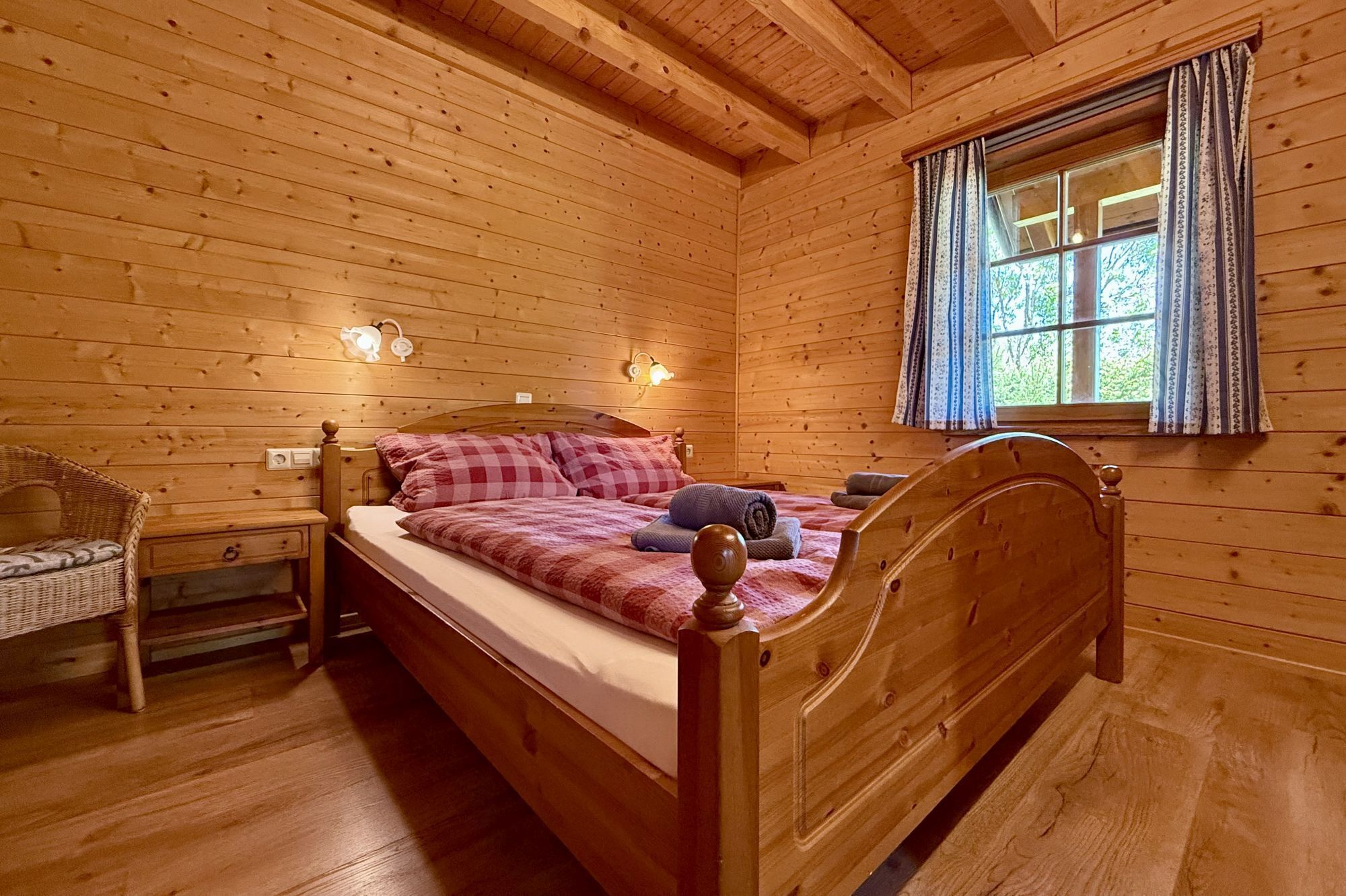 Un dormitorio de cabaña de madera con una cama, una mesita de noche, una silla y una ventana con cortinas.