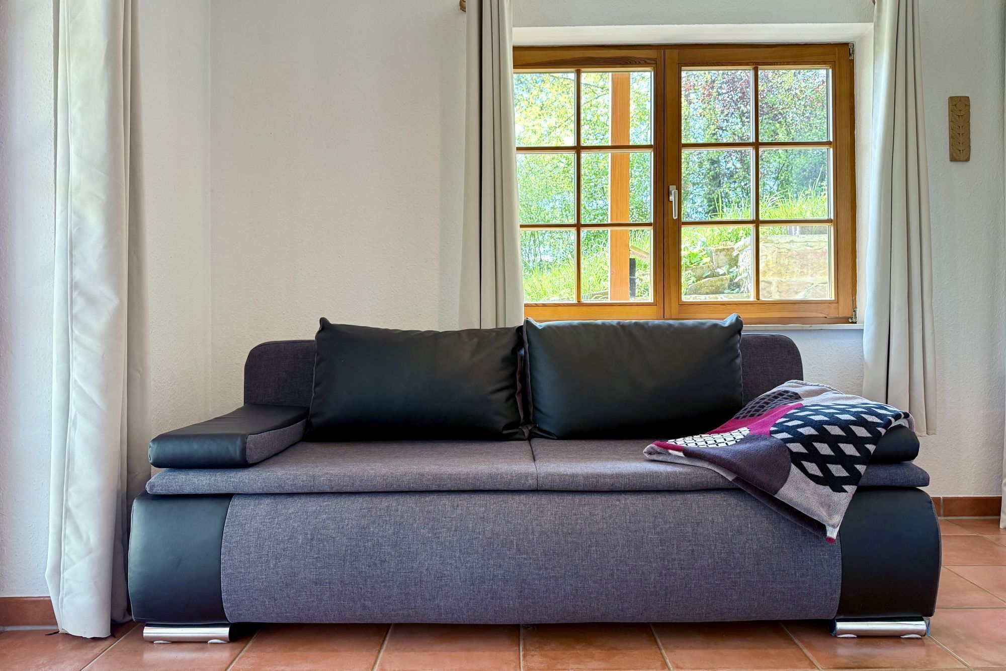 Ein graues Sofa mit zwei schwarzen Kissen und einer gemusterten Decke, vor einem Fenster mit Blick auf Grünflächen.