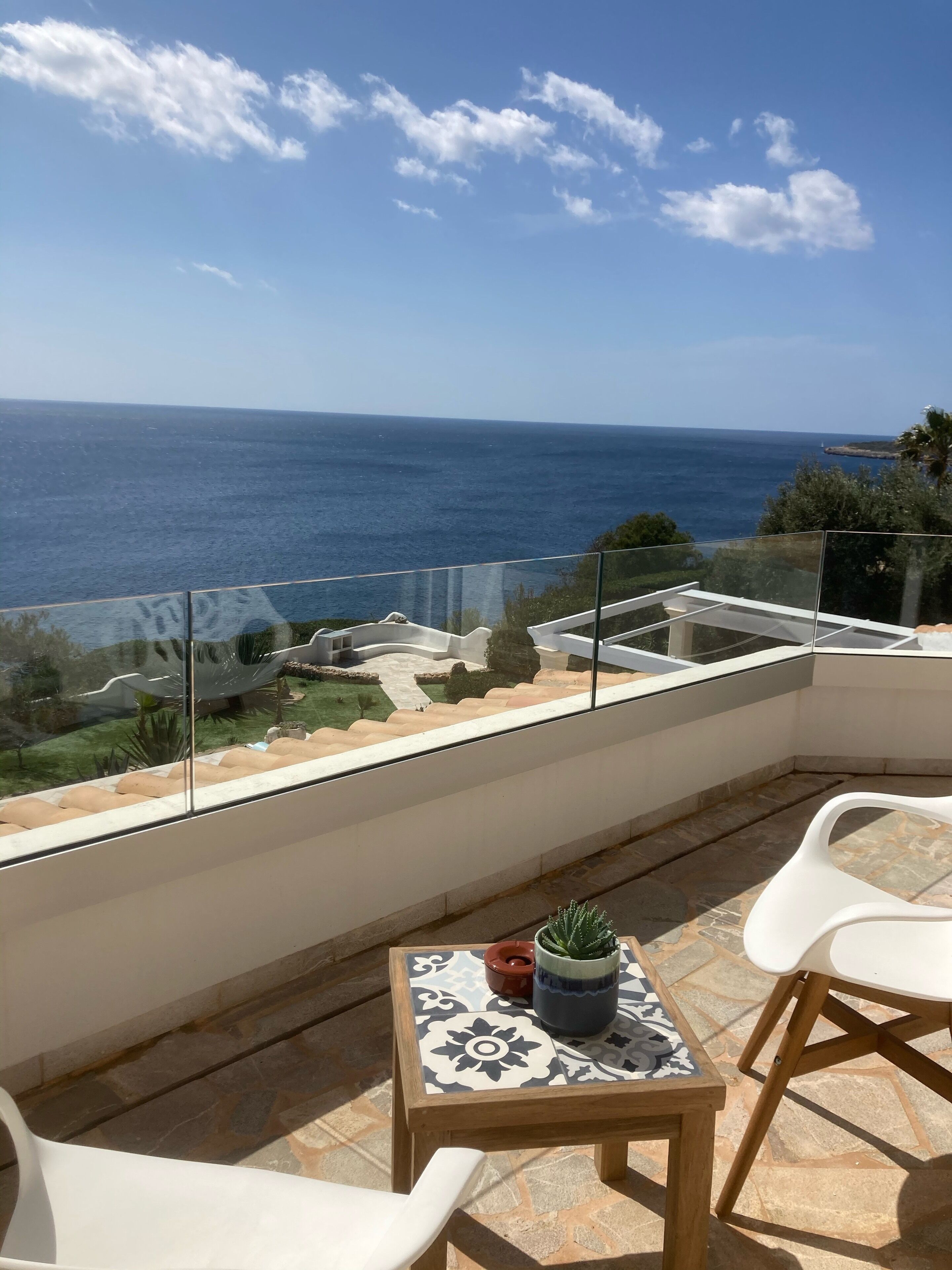 A balcony with a view of the sea, a small table with a potted plant, and white chairs.