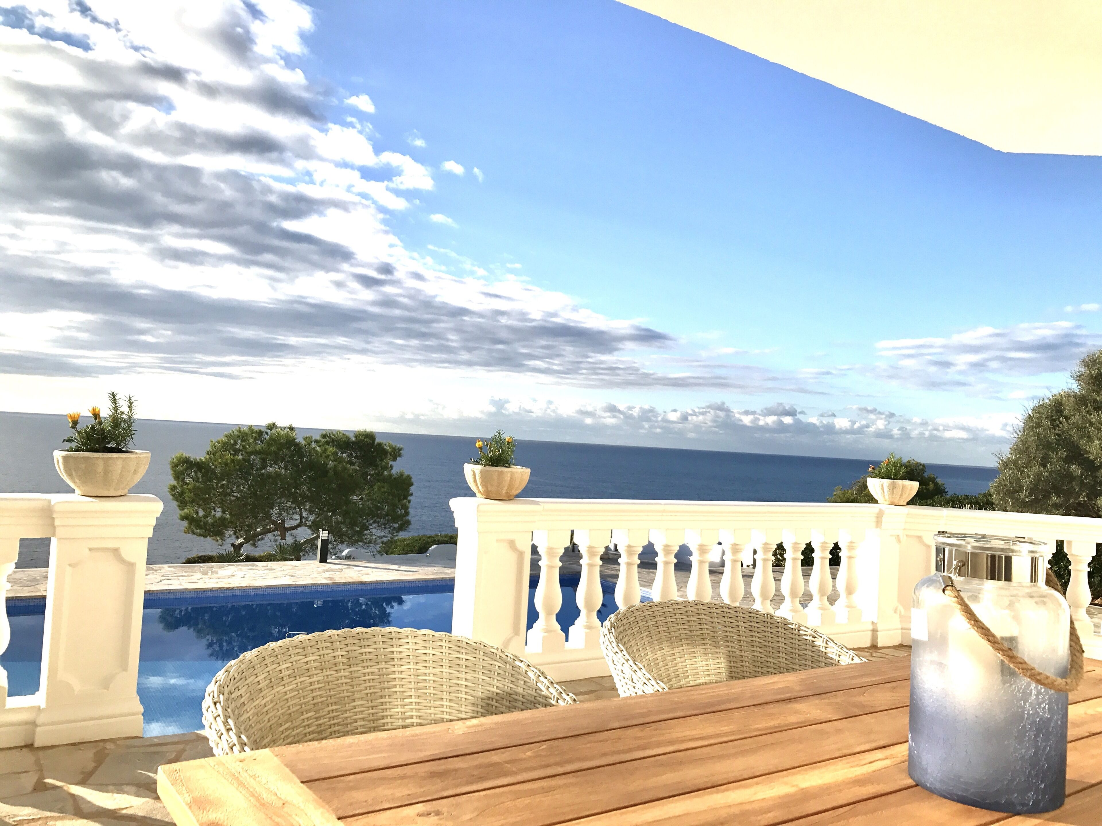 Una mesa de madera con sillas de mimbre, una piscina y vistas al mar.