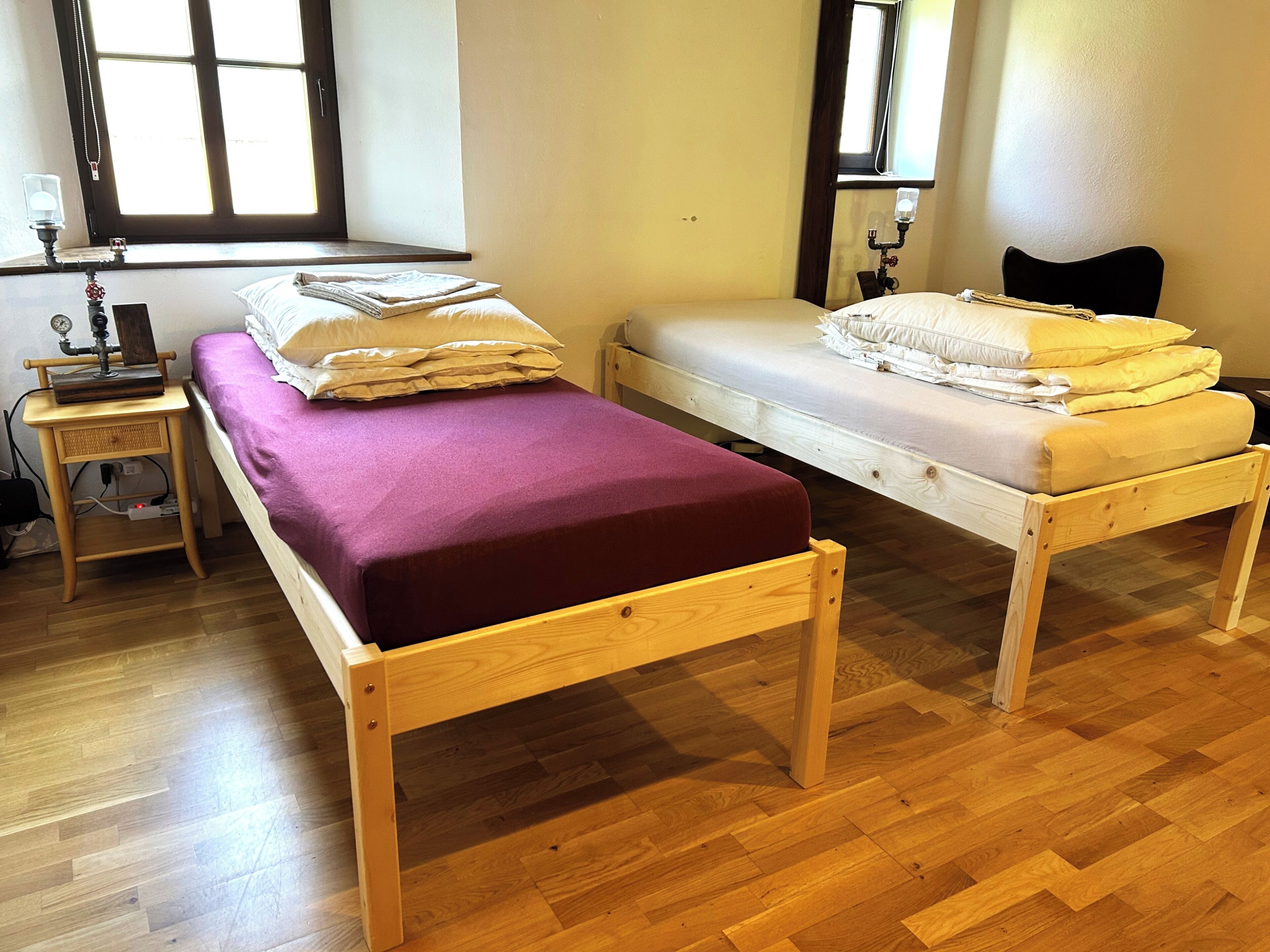 Ein Zimmer mit zwei Holzbett, einem kleinen Tisch mit Lampe und einem Fenster, das natürliches Licht hereinlässt.