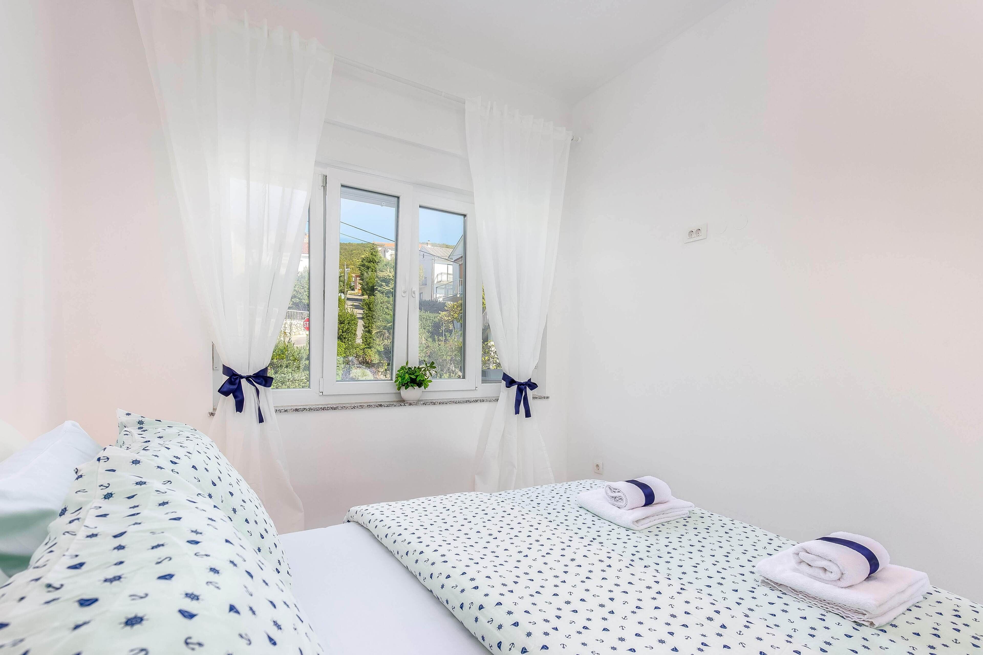 Une chambre avec deux lits, une fenêtre avec des rideaux blancs, une plante en pot et une vue sur la verdure à l’extérieur.