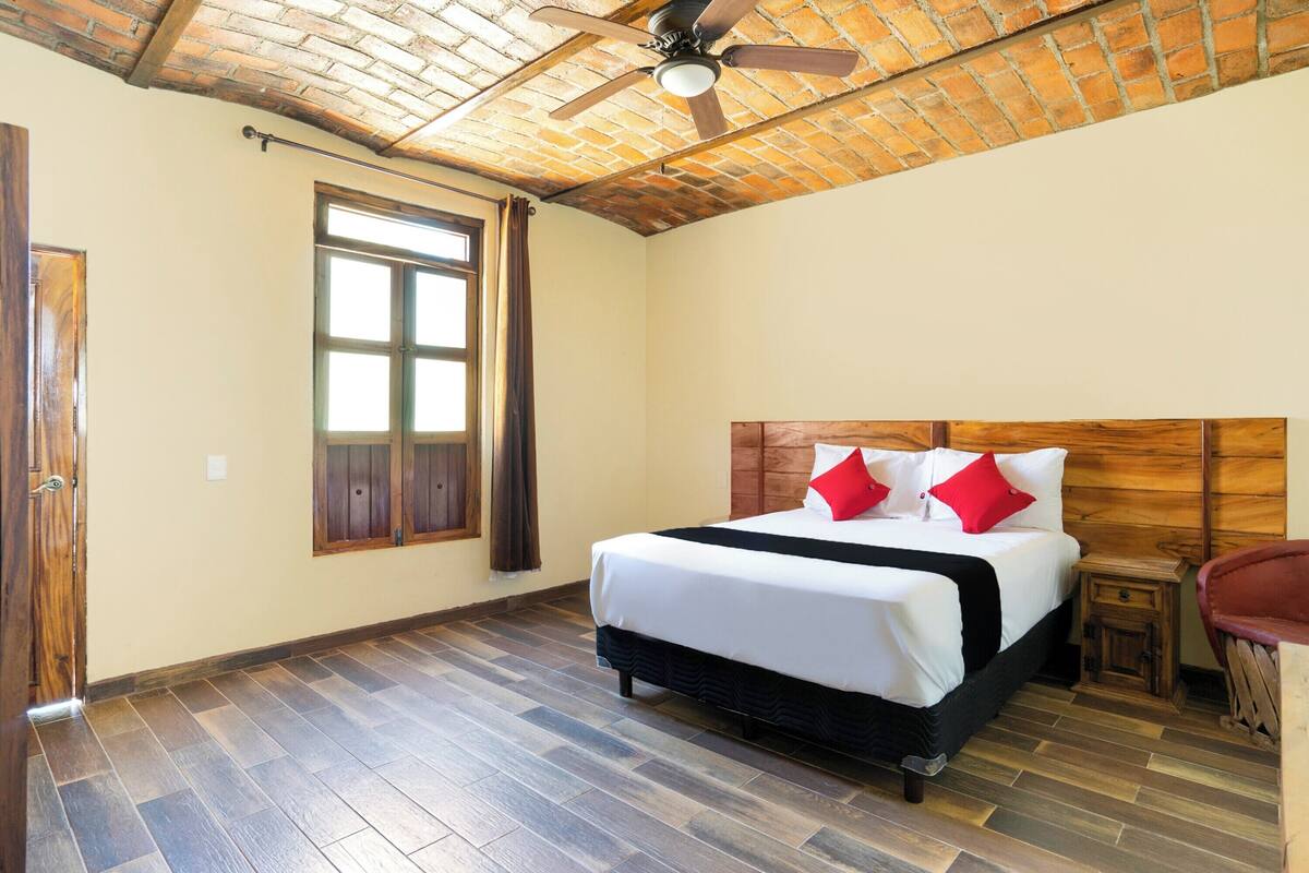 A hotel room with a wooden ceiling, a large bed with white bedding and red pillows, a small bedside table, and a window with curtains.