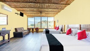 A hotel room with a brick ceiling, large windows with a mountain view, a sofa, a desk, and two beds with red pillows. - Collection O Posada Terraza (Tequila)