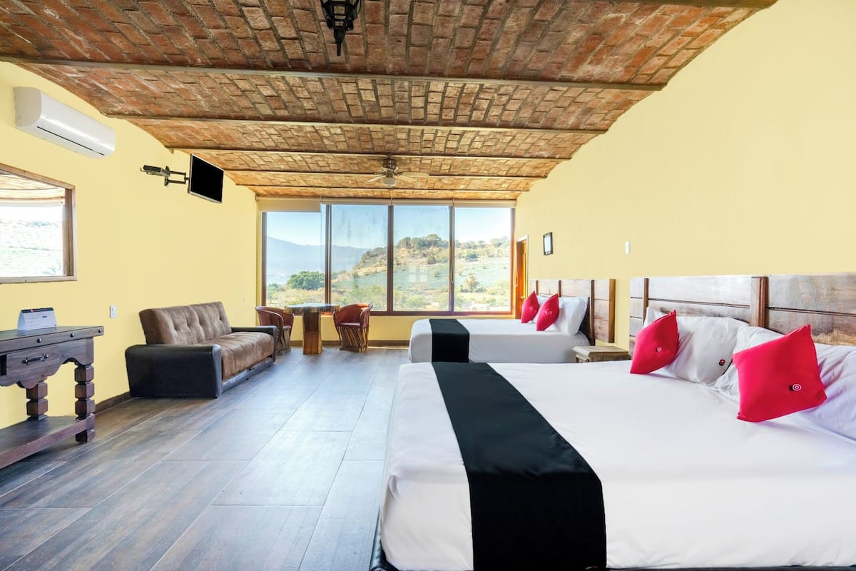 A hotel room with a brick ceiling, large windows with a mountain view, a sofa, a desk, and two beds with red pillows.