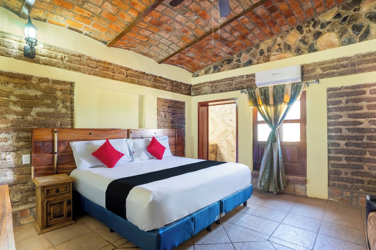 A bedroom with a brick ceiling, a wooden headboard, a bed with white bedding and red pillows, a nightstand, and a window with curtains.