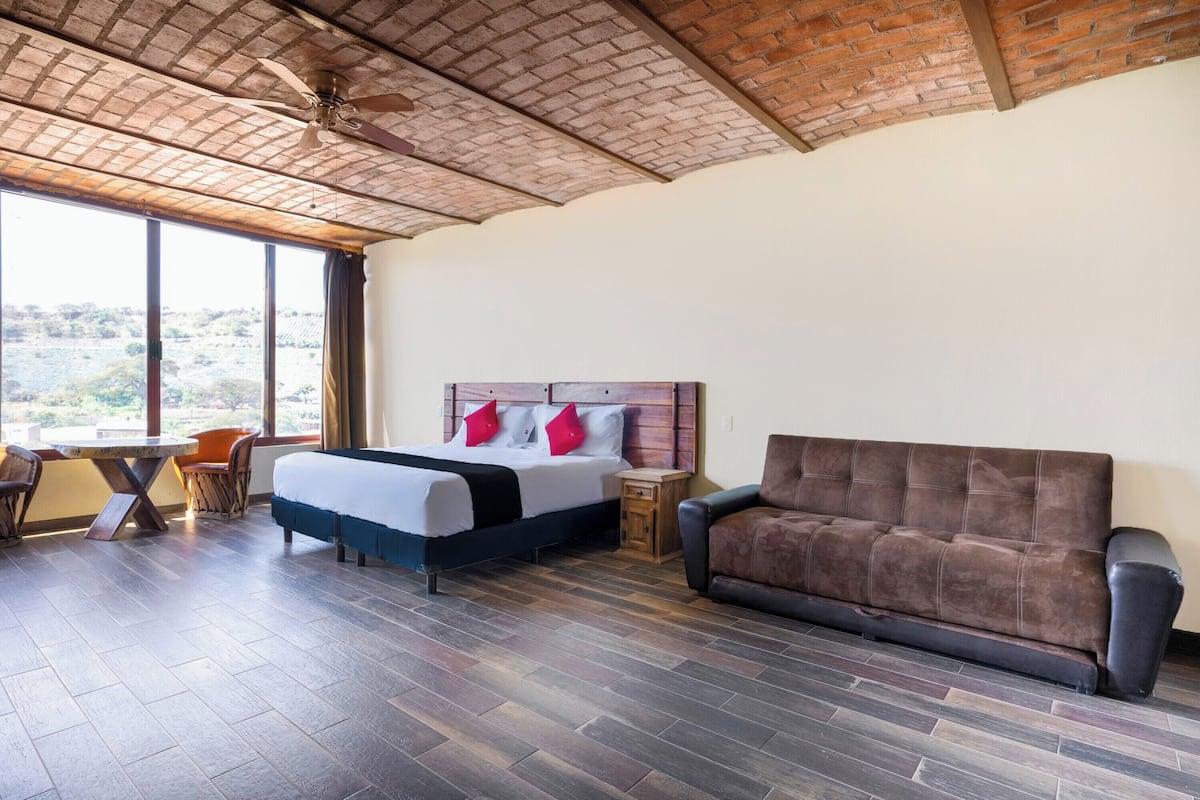 A bedroom with a bed, a sofa, a small table, and a chair. There is a window with a view of the outdoors.