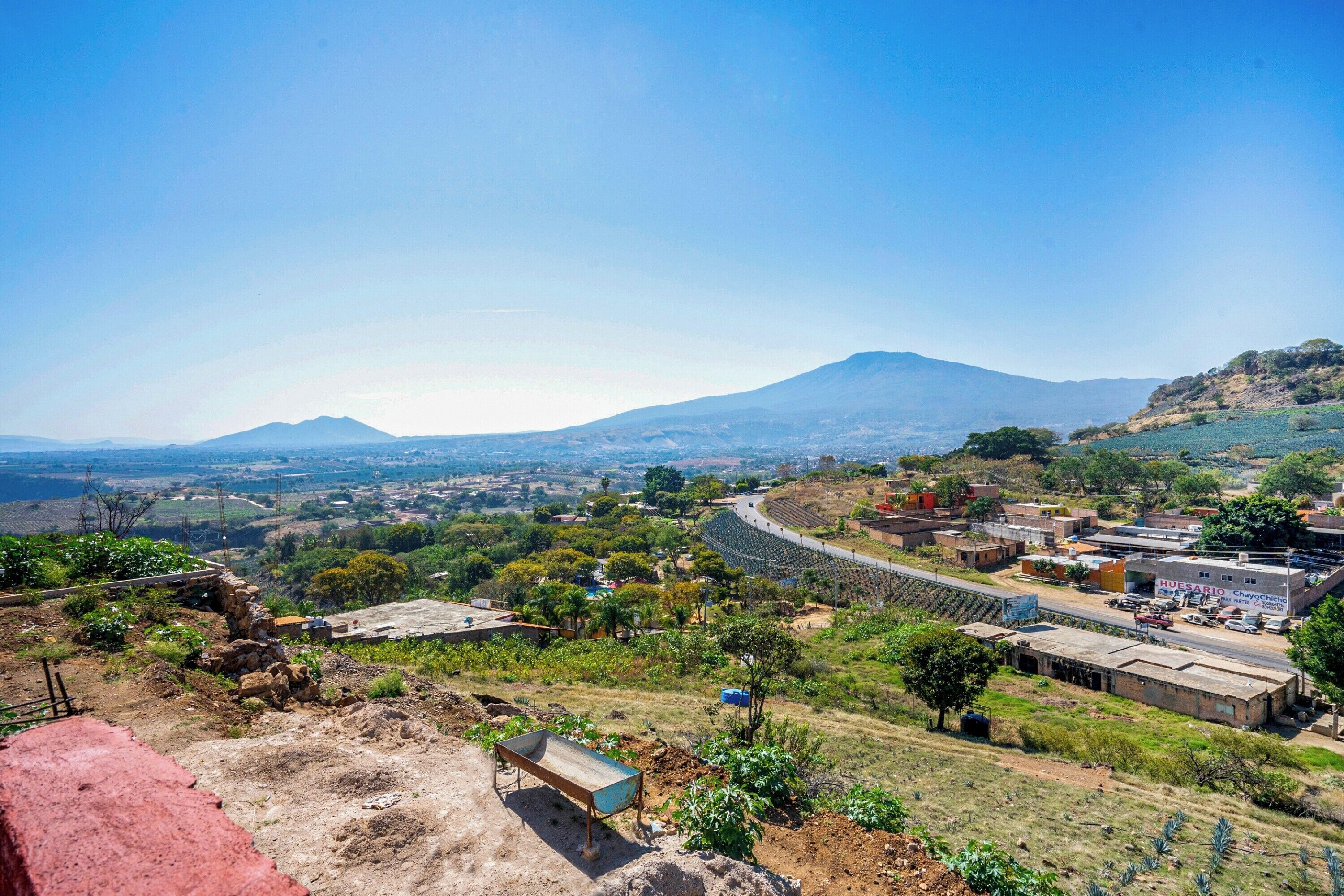 Paisaje rural con una montaña a lo lejos, un camino sinuoso y edificios dispersos.