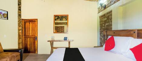 A hotel room with a bed, a wooden door, a small table with a chair, and a stone wall.
