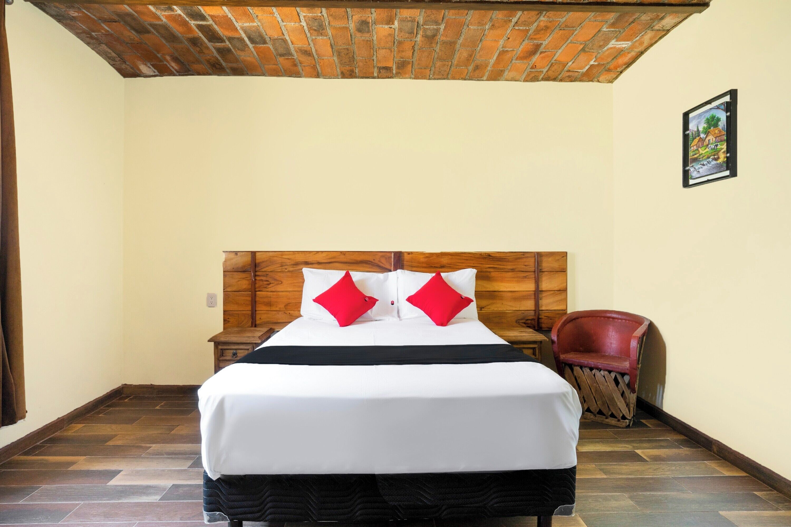 A neatly made bed with white linens and red pillows, a wooden headboard, and a red chair in a room with tiled flooring and a brick ceiling.