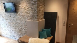 A modern room with a stone wall, a bench with teal cushions, a white fluffy footrest, and a television mounted on the wall. - Appartements Schweiberer, exclusive apartment Hochfügen (Ried im Zillertal)