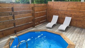Una piscina exterior con muelle de madera, escalera metálica y dos sillas reclinables.