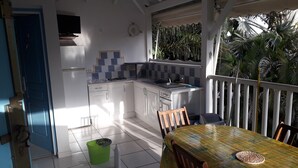 A kitchen with white cabinets, a tiled backsplash, and a small island. There is a dining table with chairs and a green trash bin. - ti case gwada bungalow rentals "ti poisson" (Sainte-Anne)