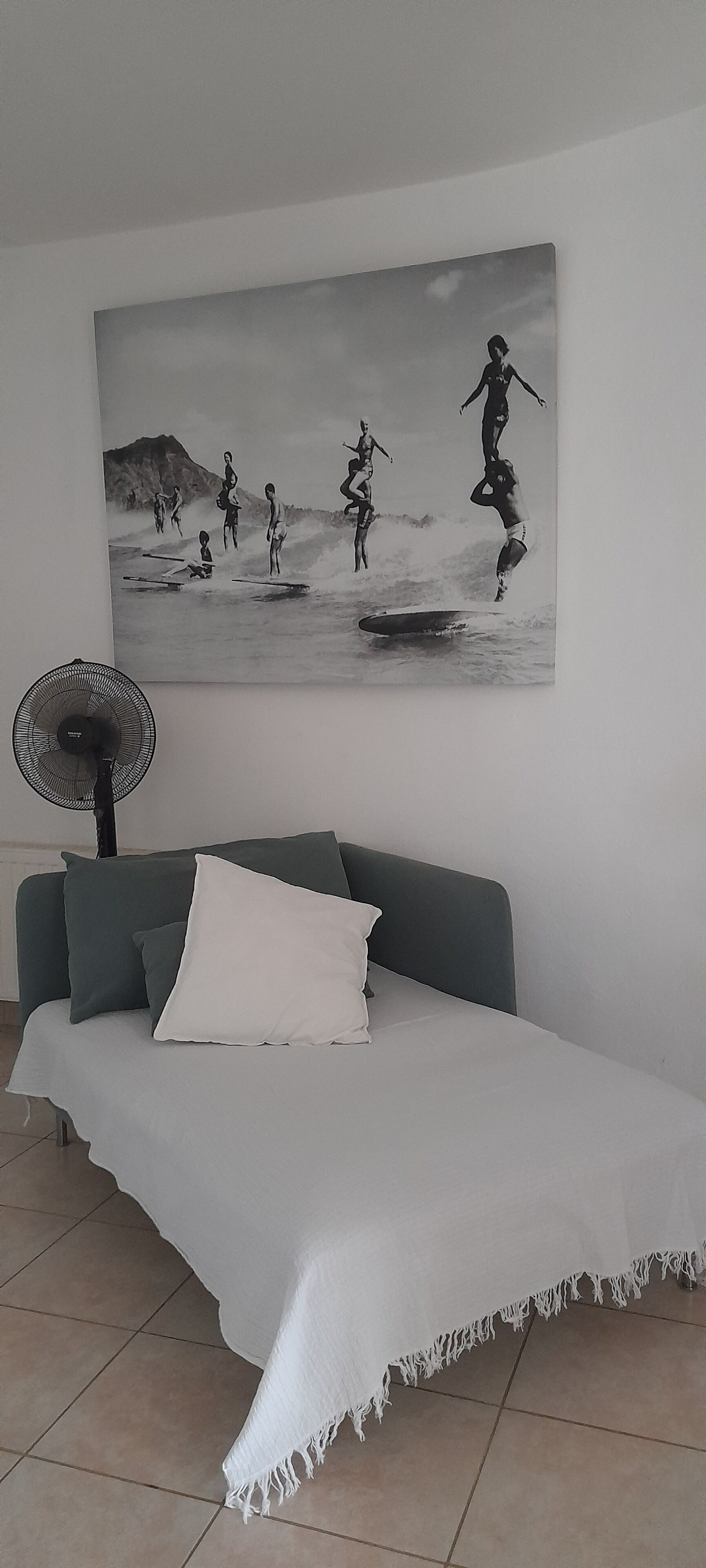 A bedroom with a bed, a wall-mounted fan, and a large beach-themed artwork.