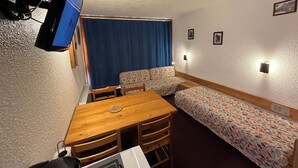 Una habitación compacta con una mesa de madera, dos sofás estampados, una cortina azul y un televisor de pared.